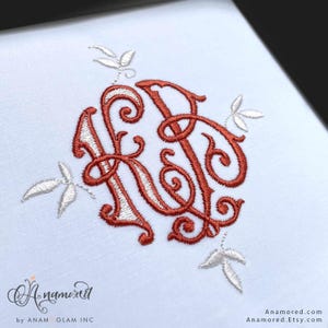 May include: White fabric with an embroidered monogram in a reddish-brown color, featuring the letters "KR" intertwined with decorative swirls. Delicate white leaf accents surround the monogram. The text "Anamored" and "Anamored.Etsy.com" are also visible.