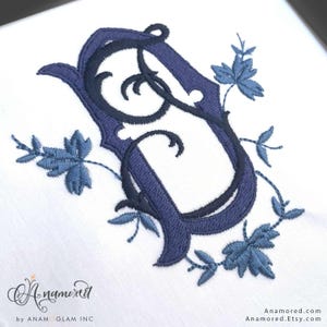 May include: Embroidered monogram letter "J" in navy blue with black accents and light blue leaf accents. The design is on a white background. The Etsy shop name "Anamored" is in the bottom left corner.