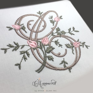 May include: Embroidered design of a stylised letter "S" with pink roses and green leaves. The design is surrounded by a decorative border. The text "Anamored" is written below the design.