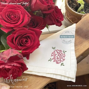 May include: A white linen napkin featuring a pink embroidered monogram with green leaf accents. The napkin is positioned next to a bouquet of deep red roses. A circular stamp on the fabric reads 100% HUMAN MADE NO AI USED.
