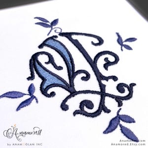 Op de afbeelding: Geborduurde monogramletter "A" in marineblauw en lichtblauw, omringd door gestileerde bladeren. Het ontwerp staat op een witte achtergrond, met de Etsy shopnaam "Anamored" linksonder.