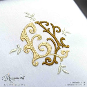May include: A gold and cream embroidered monogram with a decorative floral design. The monogram is in a circle shape and the letters are intertwined.