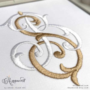 May include: An embroidered monogram featuring the intertwined letters S and D, rendered in metallic gold and white thread on a white fabric background. The design displays ornate, swirling script calligraphy with a dense, textured stitch pattern.