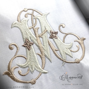 Może przedstawiać: Haftowany monogram litery "M" w kolorze białym i beżowym z ozdobnymi detalami kwiatowymi. Monogram znajduje się na białym tle tkaniny.