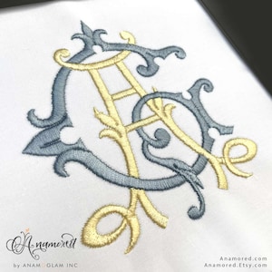 May include: A white fabric surface featuring an intricate embroidered monogram of the letters A and G. The letter A is stitched in a pale yellow thread, while the letter G is rendered in a slate blue thread with decorative flourishes.