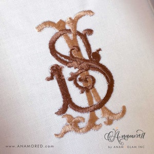 Könnte beinhalten: Ein Monogramm, das auf einen weißen Stoffhintergrund gestickt ist. Das Monogramm ist in braunem und goldenem Garn gestickt und zeigt die Buchstaben "S" und "U", die miteinander verschlungen sind.