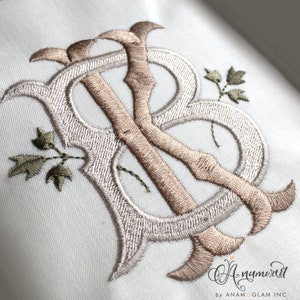 May include: Embroidered monogram with the letters "KB" in a decorative script font. The letters are in a light brown color and are surrounded by a white outline. There are small green leaves embroidered around the letters.