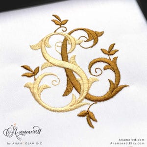 May include: Embroidered monogram featuring the letters "S" and "A" in elegant script, intertwined with floral accents. The design uses gold and light beige thread on a white background. The design is from Anamored.