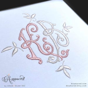May include: Close-up of a white fabric with an embroidered monogram featuring the letters "K" and "R" in a delicate, swirling script. The "K" is in a soft pink, while the "R" is in a light beige. Small embroidered leaves surround the letters.