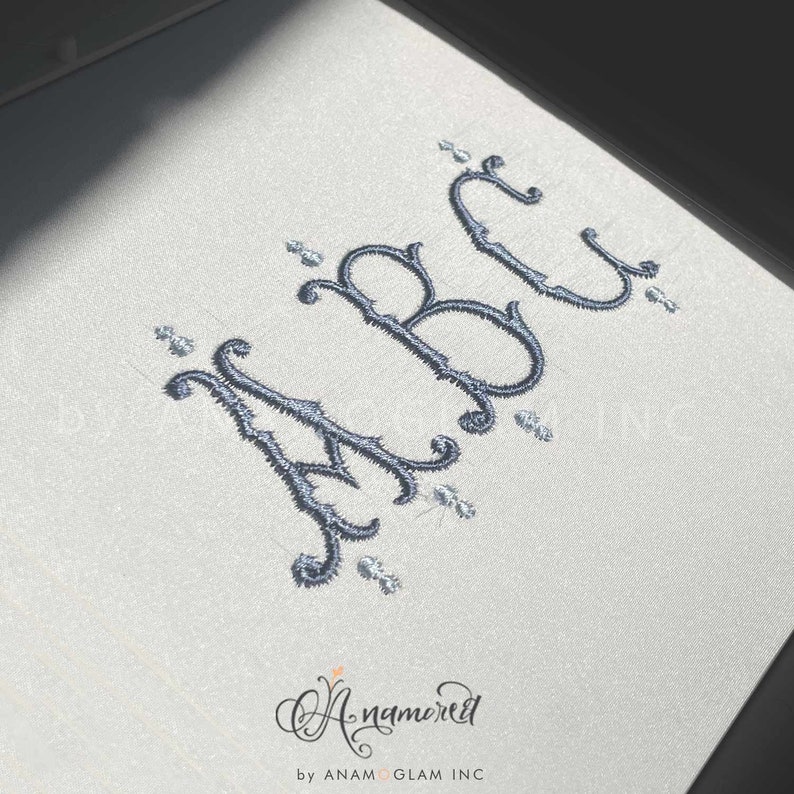 May include: Embroidered letters "ABC" in a decorative script font with silver embellishments on a white background. The letters are in a cursive style with flourishes and swirls.