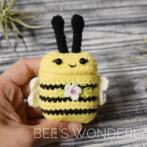 May include: A crocheted bee-shaped phone case with black antennae, yellow and black stripes, and a white flower with green leaves. The case is held in a hand.