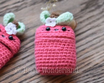 May include: A pink crocheted Airpods case with a green leaf and white flower on top. The case has a cute cartoon face with black eyes and a pink nose.