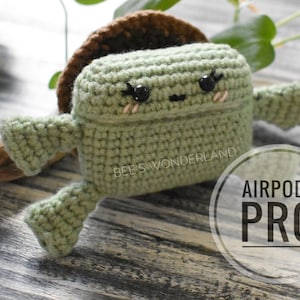 May include: A green crocheted Airpods Pro case with a brown crocheted shell on top. The case has a cute face with black eyes and pink cheeks. The text "AIRPODS PRO" is written in white on a black circle.