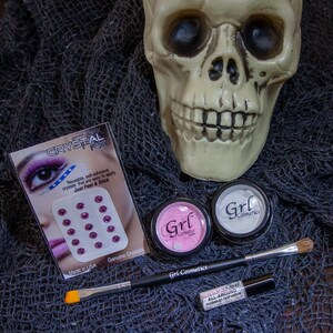 May include: A skull figurine, two small jars of pink and white makeup, a makeup brush, and a small tube of makeup adhesive. The jars have the text "Grl Cosmetics" on them. The makeup adhesive tube has the text "All-Around Makeup Adhesive". The skull is on a black mesh fabric. The packaging for a set of self-adhesive crystals is visible in the background. The packaging has the text "Crystal FX" and "Just Peel & Stick".