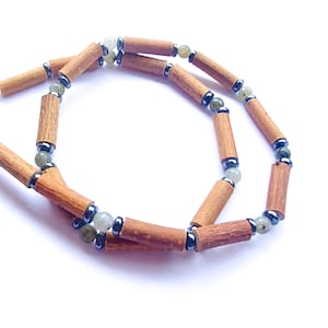Puede incluir: Un collar con cuentas cilíndricas de madera, cuentas verde claro y gris oscuro. Las cuentas de madera son de color marrón claro. El collar tiene un cierre plateado. Las cuentas se miden en centímetros.