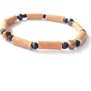 May include: A bracelet with light brown wooden beads, black lava beads, and tiger's eye beads. The bracelet is presented on a white background. The wooden beads are cylindrical, and the tiger's eye beads are round. The bracelet's design highlights the natural materials.