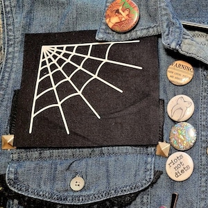 Może przedstawiać: Jeansowa kamizelka z czarnymi kieszeniami z białymi wzorami pajęczyn. Kamizelka jest ozdobiona różnymi przypinkami, w tym przypinką w kształcie serca ze słowem "cunt" i innymi elementami dekoracyjnymi.