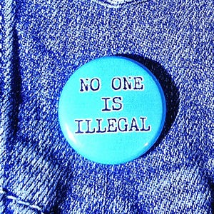 Może przedstawiać: Jasnoniebieska przypinka z białym napisem "NO ONE IS ILLEGAL". Przypinka jest przypięta do kieszeni niebieskiej kurtki dżinsowej, z widocznymi szwami i guzikiem.