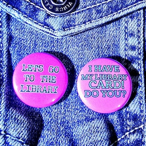 May include: Two pink circular badges with white text on a blue denim background. One badge reads "LETS GO TO THE LIBRARY" and the other says "I HAVE MY LIBRARY CARD! DO YOU?" A silver button and a blue badge are also visible.