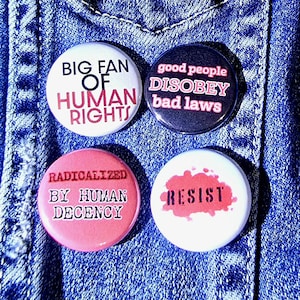 May include: Four round buttons with text on a blue denim background. The buttons have the following text: "BIG FAN OF HUMAN RIGHTS", "good people DISOBEY bad laws", "RADICALIZED BY HUMAN DECENCY", and "RESIST".