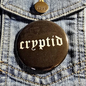 May include: A black button with the word "cryptid" in white gothic lettering, pinned to a blue denim jacket.