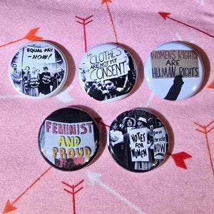 May include: Five circular feminist badges with different slogans and images. The badges display phrases such as "Equal Pay - Now!", "My Clothes Are Not My Consent", and "Feminist and Proud". The badges are set against a pink background with arrow patterns.