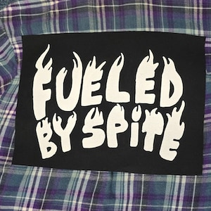 Könnte beinhalten: Ein blaues und violettes kariertes Flanellhemd mit einem schwarzen Patch, der auf der Rückseite aufgenäht ist. Der Patch hat weißen Text, der "Fueled by Spite" lautet, mit Flammen um den Text herum.