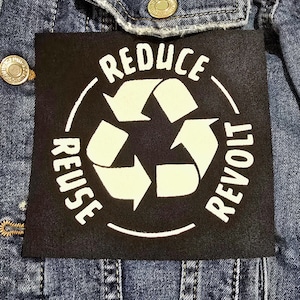 May include: A denim jacket with a black patch featuring a recycling symbol and the words "Reduce, Reuse, Revolt." Two round pins are also visible, one with the text "No War But Class War" and another with the text "When Injustice Becomes Law Resistance Becomes Duty."