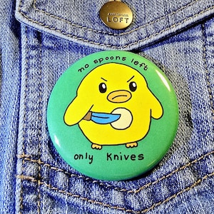May include: A green and yellow button with a cartoon chick holding a knife. The text on the button reads "no spoons left only knives".