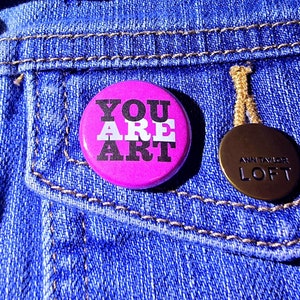 May include: A pink button with the words "YOU ARE ART" in black and white lettering on a denim background.