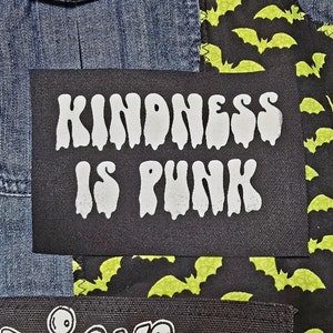 May include: A black patch with white dripping text that reads "Kindness is Punk" sewn onto a denim jacket with a black and green fabric with a bat pattern.