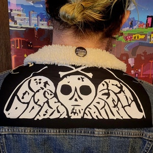 May include: A black denim jacket with a white skull and crossbones patch on the back. The patch features a skull with wings and the text "Death Before Dishonor". The jacket also has a white sherpa collar and a small black button with the text "Friendly Neighbourhood Anarchist".