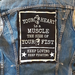 Könnte beinhalten: Ein schwarz-weißes Patch, das auf eine Jeansjacke genäht ist. Das Patch zeigt ein Herz mit einer Faust und den Text "YOUR HEART is a MUSCLE THE SIZE OF YOUR FIST KEEP LOVING KEEP FIGHTING".