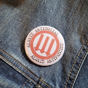 May include: A white button with red text and a red symbol. The text reads "Always Antisocial Always Anti-Fascist". The symbol is three arrows pointing to the left.