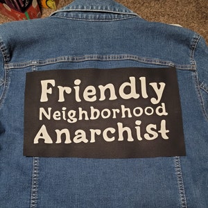 May include: A blue denim jacket with a black patch sewn on the back. The patch has white text that reads "Friendly Neighborhood Anarchist".