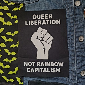 May include: A black patch with white text that reads "Queer Liberation" and "Not Rainbow Capitalism". A white raised fist is in the center of the patch.
