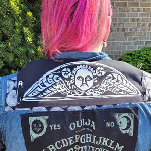 Peut inclure: Une personne portant une veste en jean avec un patch noir représentant un ange ailé et les mots "Ouija Mystic Oracle". La veste a également un patch avec les lettres du planchette.
