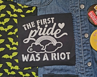 May include: A black patch with white text that reads "The first pride was a riot". The patch has a rainbow and a heart above the text. The patch is sewn onto a denim jacket with other patches.