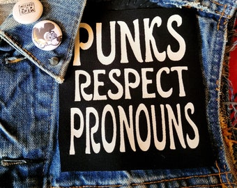 May include: A black denim jacket with a white patch that says "Punks Respect Pronouns". The jacket has several pins, including one with a cartoon character.