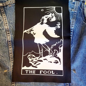 May include: Black and white tarot card patch featuring an image of a figure walking towards a sun with a white background. The text "THE FOOL." is printed at the bottom of the card.