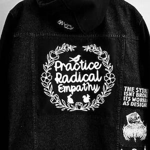 May include: Black denim jacket with a white embroidered design on the back that says "Practice Radical Empathy" in a wreath. The jacket also has other patches with text and designs.