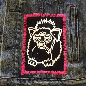 May include: A black and white embroidered patch of a cartoon furry creature with a cigarette in its mouth. The patch is sewn onto a blue denim jacket with pink stitching around the edges.