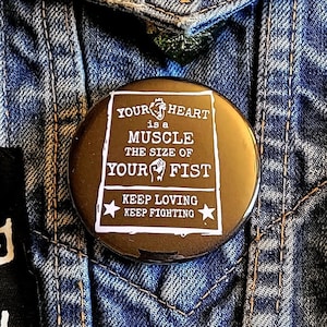 May include: A black and white button with the text "YOUR HEART is a MUSCLE THE SIZE OF YOUR FIST KEEP LOVING KEEP FIGHTING" on a denim jacket.