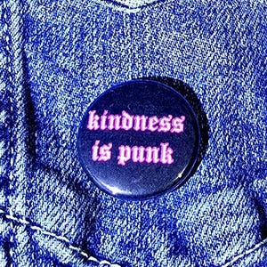 Może przedstawiać: Okrągła, czarna przypinka z różowym napisem "kindness is punk". Przypinka jest przypięta do kieszeni niebieskiej kurtki dżinsowej, z widocznymi białymi szwami i teksturowanym wyglądem.