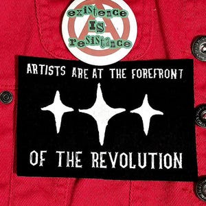May include: A red jacket with black buttons and patches. Patches include a pin that says "Friendly Neighborhood Anarchist", a round pin with "existence IS resistance", and a black patch with "ARTISTS ARE AT THE FOREFRONT OF THE REVOLUTION".