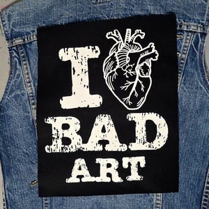 Può includere: Toppa nera con testo bianco e un grafico di cuore anatomico bianco. Il testo dice "I ♥ BAD ART".