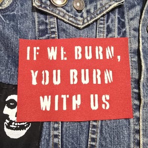 May include: Close-up of a denim jacket with patches and buttons. A red rectangular patch with the white text "IF WE BURN, YOU BURN WITH US" is prominently displayed. A black and white skull patch is also visible.