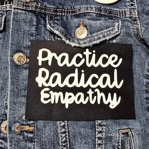 Peut inclure: Une veste en jean noire avec un patch noir cousu sur le devant. Le patch a un texte blanc qui dit "Practice Radical Empathy".
