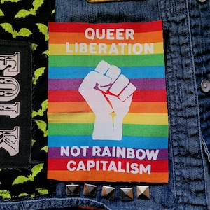 May include: A rainbow-coloured patch with a raised fist in the centre. The text "Queer Liberation Not Rainbow Capitalism" is printed on the patch.