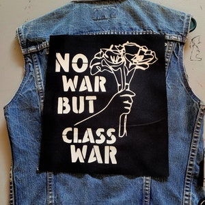 May include: A black patch with white text and a graphic of a hand holding flowers. The text reads "No War But Class War".